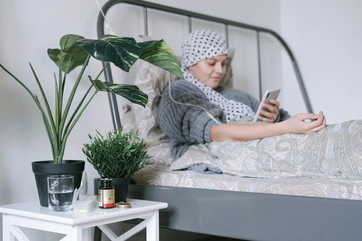 A woman undergoing cancer treatment relaxes in bed using her phone, surrounded by plants.